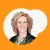 Professional headshot of Tamara Rabenold, CEO of Vaudra International, smiling warmly. She has curly blonde hair, wears a black leather jacket over a patterned blouse, and is set against an orange background with a white abstract shape.