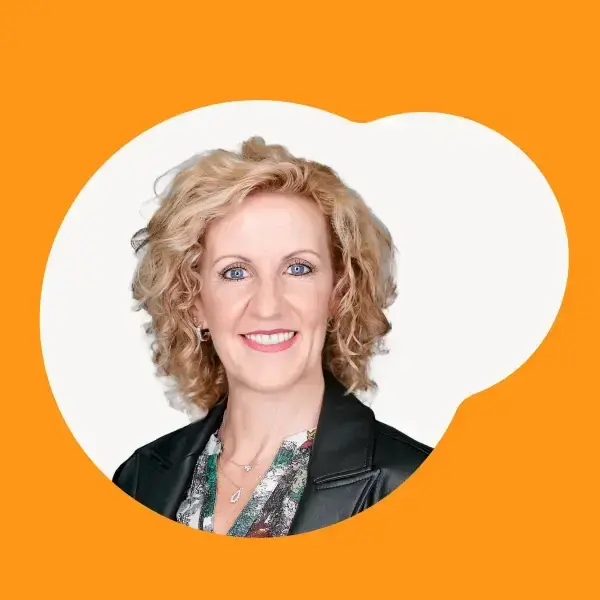 Professional headshot of Tamara Rabenold, CEO of Vaudra International, smiling warmly. She has curly blonde hair, wears a black leather jacket over a patterned blouse, and is set against an orange background with a white abstract shape.